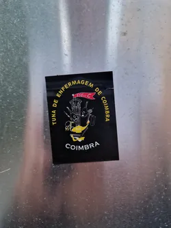 Black sticker with the logo of Tuna de Enfermagem de Coimbra. It features a clock tower illustration and text in Portuguese. 
