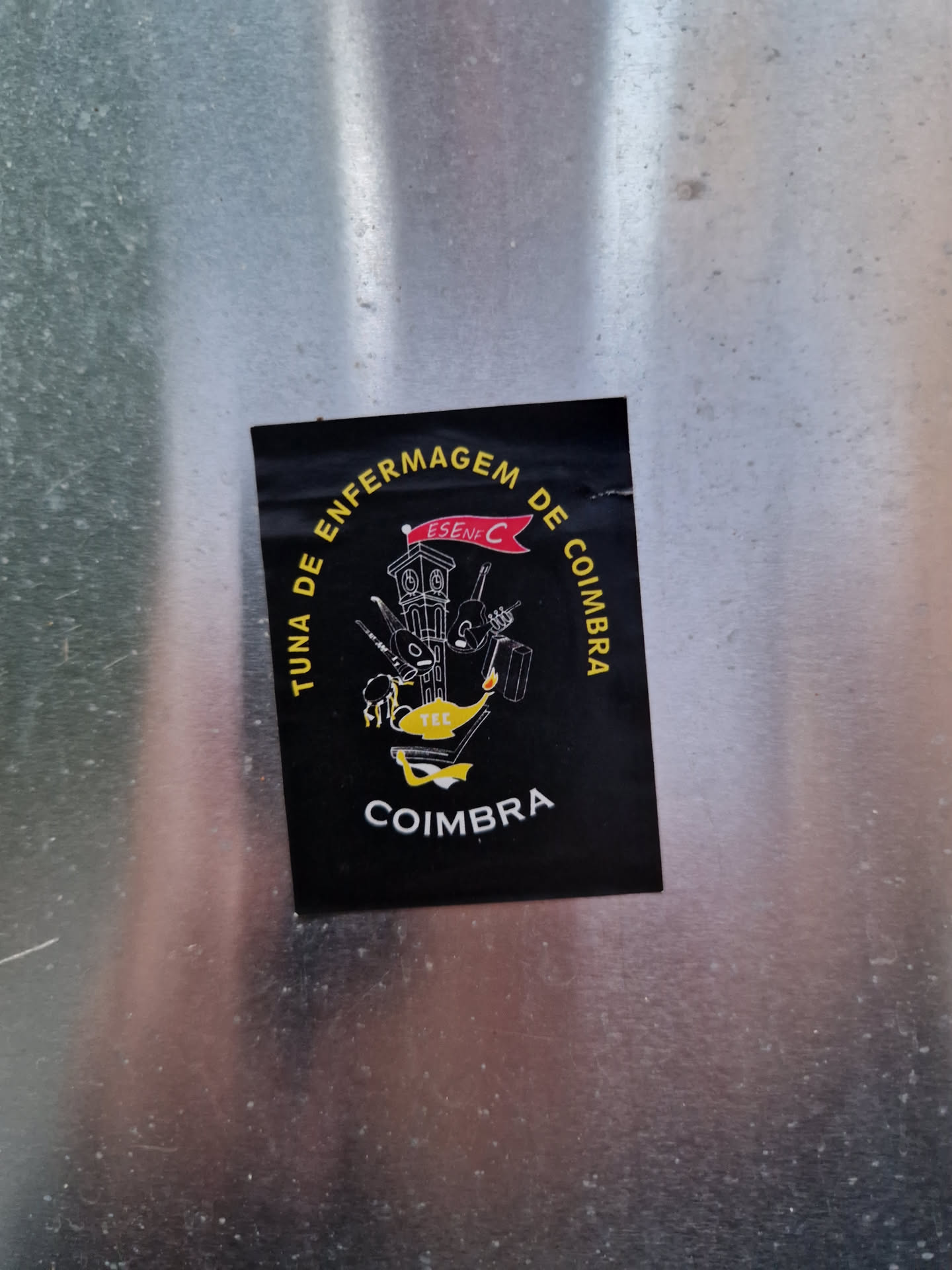 Black sticker with the logo of Tuna de Enfermagem de Coimbra. It features a clock tower illustration and text in Portuguese. 