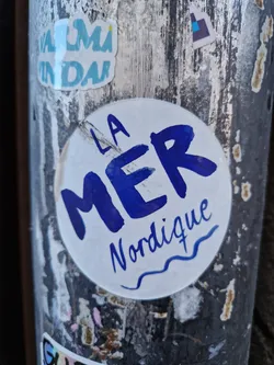 Round sticker with the text "LA MER Nordique" written in blue.  The words "La Mer" are larger and bolder than "Nordique", which is written in a smaller, more cursive font. A small wavy line is under "Nordique".