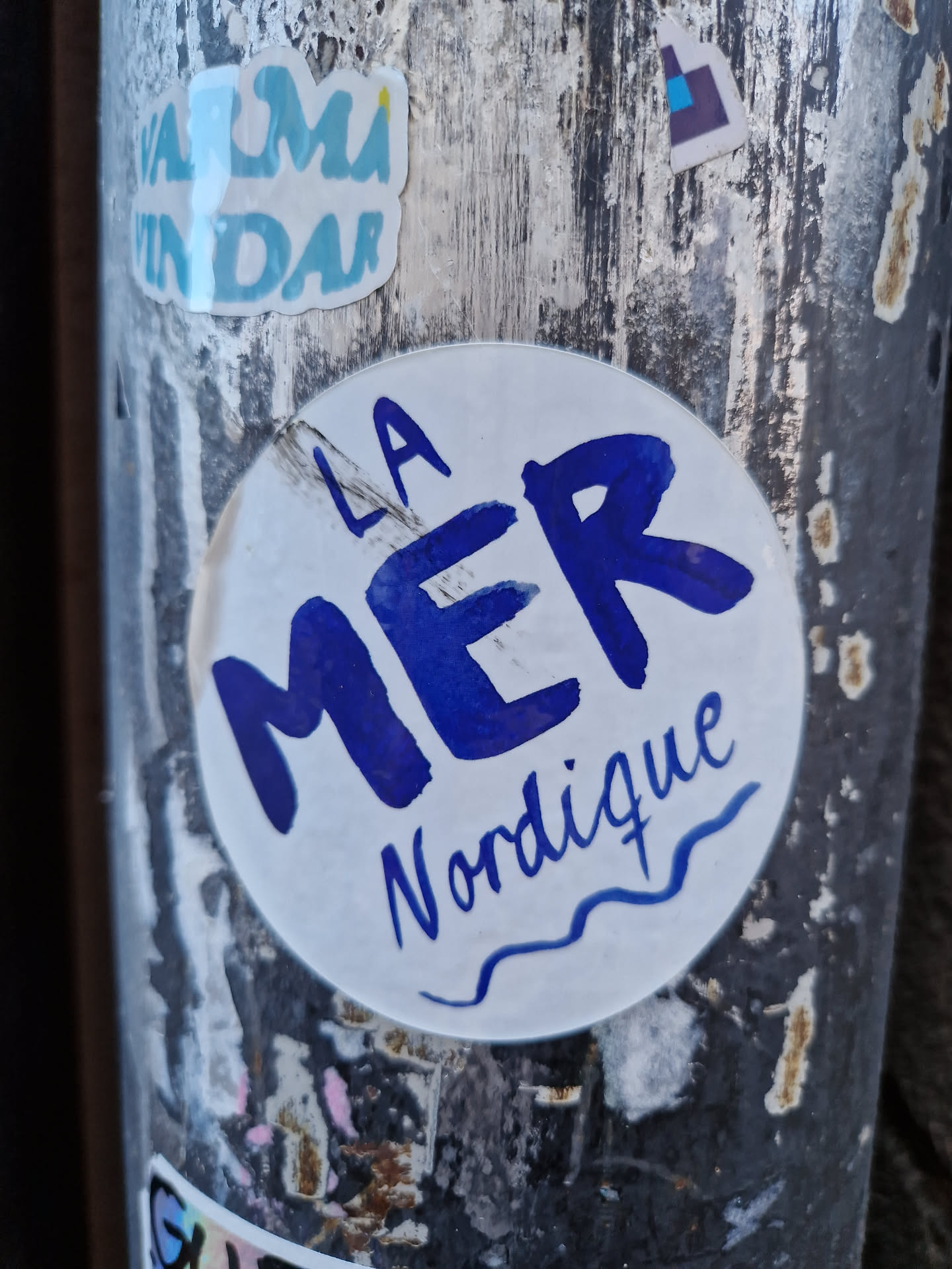 Round sticker with the text "LA MER Nordique" written in blue.  The words "La Mer" are larger and bolder than "Nordique", which is written in a smaller, more cursive font. A small wavy line is under "Nordique".