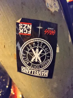 A black and white sticker with a circular design featuring a pentagram and text that says "HAVELLAND",  "M1666LUCKTE WELT", and other symbols.  There are additional texts above, including "FCK NZS" and what seems to be a stylized cross and another text.