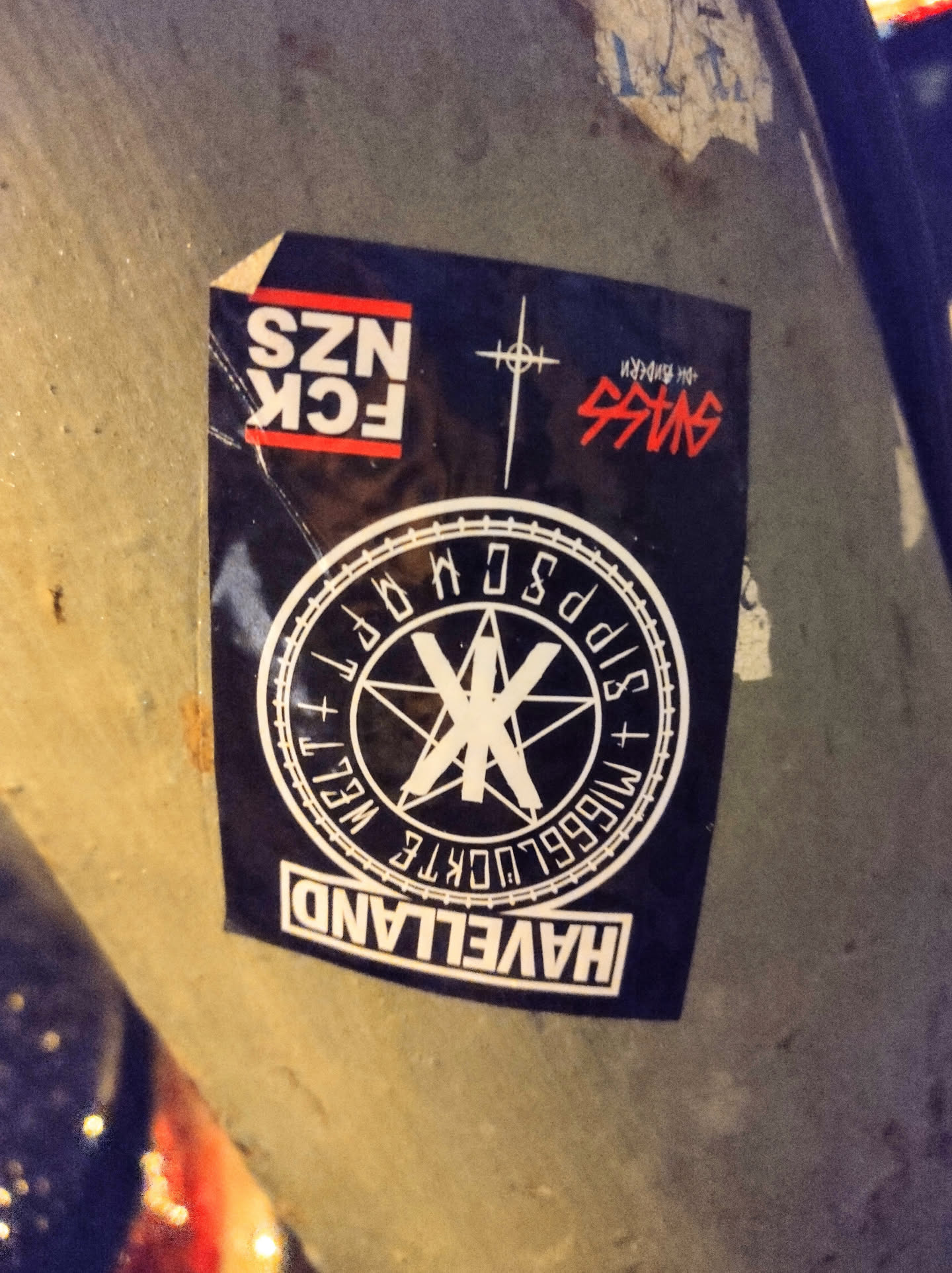A black and white sticker with a circular design featuring a pentagram and text that says "HAVELLAND",  "M1666LUCKTE WELT", and other symbols.  There are additional texts above, including "FCK NZS" and what seems to be a stylized cross and another text.