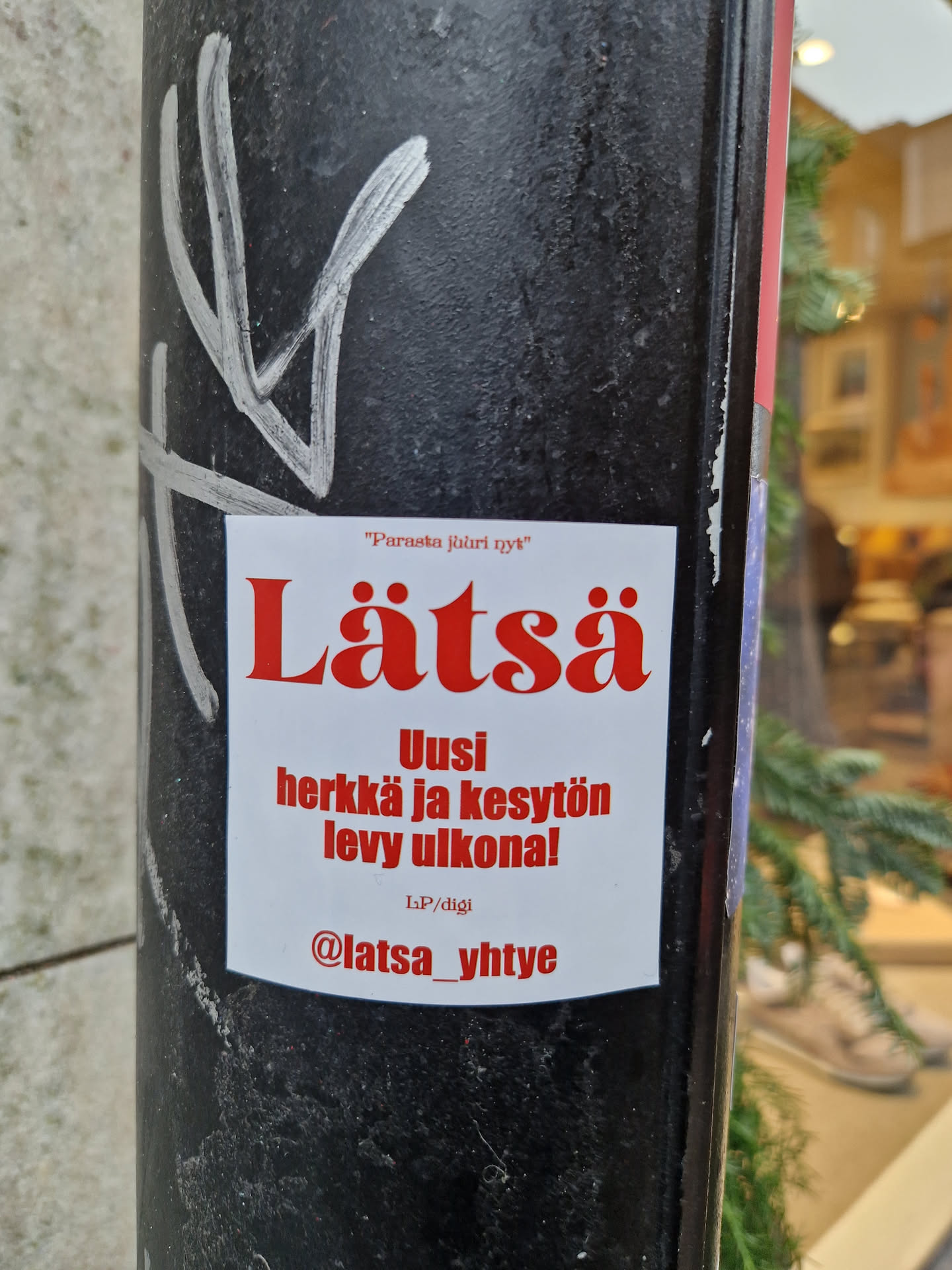 Sticker promoting the release of a new album by Lätsä. The text on the sticker says: "Uusi herkkä ja kesytön levy ulkona!" which translates to: "New sensitive and tame album outside!"  There is also a social media handle @latsa_yhtye. 