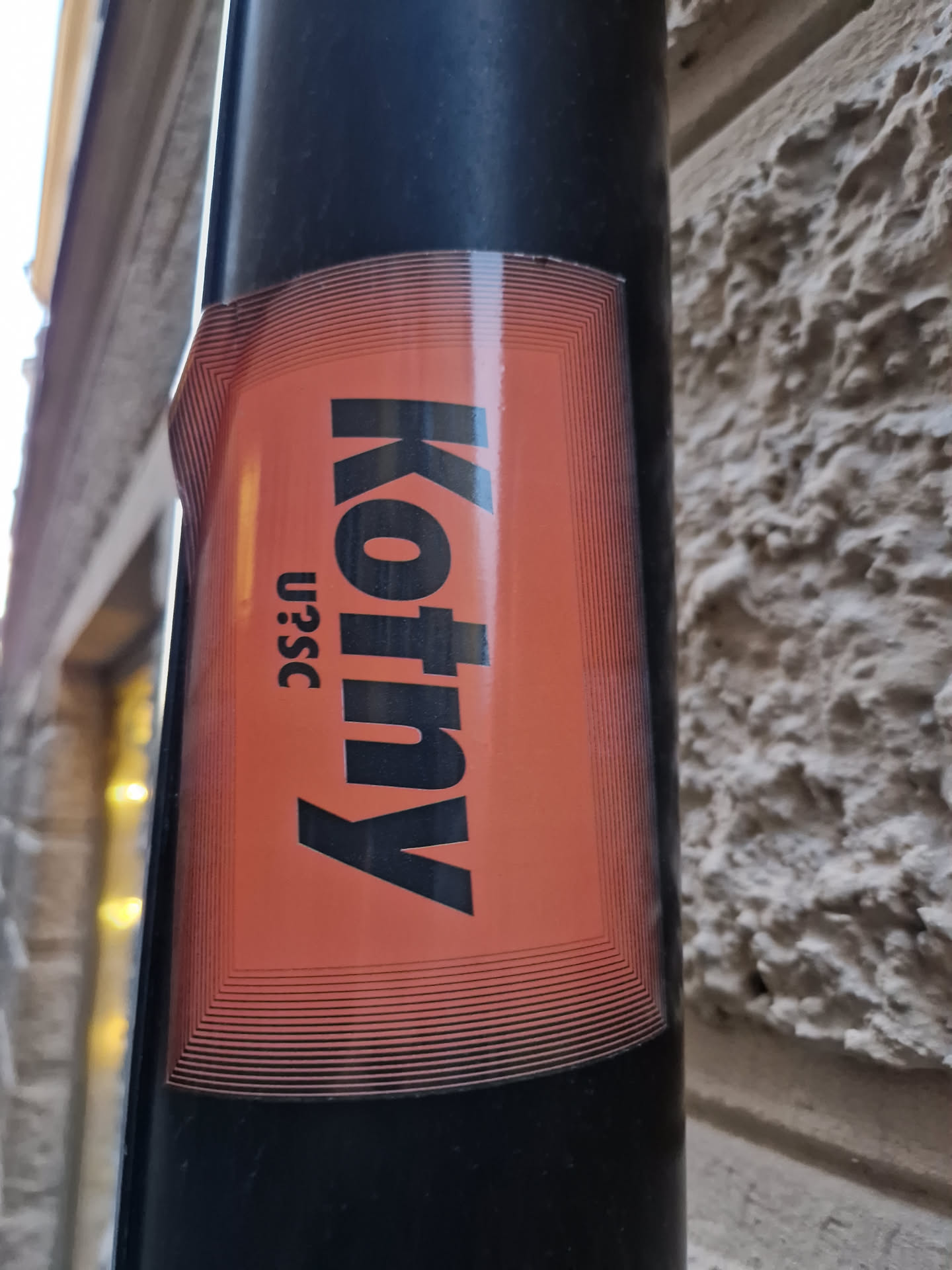 An orange sticker with the word Kotny in black text. The sticker has a decorative border of concentric lines. The text "U25C" is visible at the bottom.