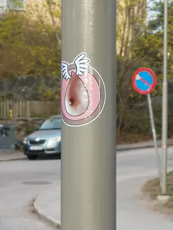 The sticker features a stylized, light brown human breast with a darker brown nipple, set against a distressed pink background within a circular frame. White, feathery wings extend from the top of the breast. The entire design is outlined in black, followed by a clean white border, giving it a cartoonish and whimsical appearance. There are some signs of wear and slight peeling on the edges.