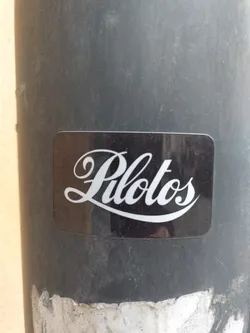 A black sticker with the word "Pilots" written in a white, stylized script font. The sticker is affixed to a dark gray pole.