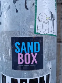A sticker for a music and entertainment company called SANDBOX. The sticker is black with the word 'SAND' in blue and 'BOX' in pink.  The text 'music & entertainment' appears below in a smaller font.