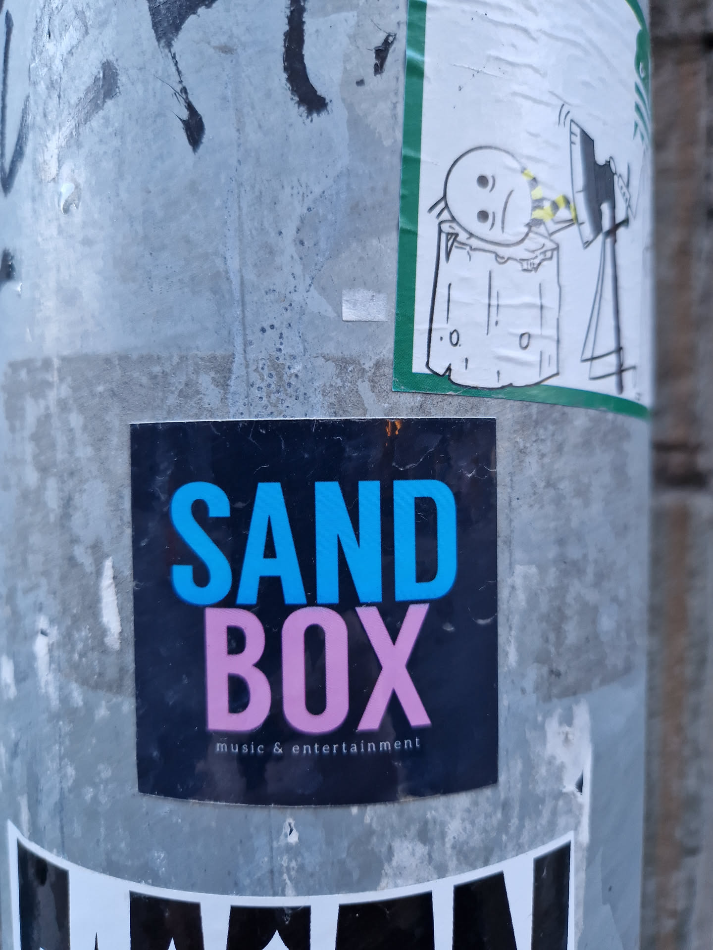 A sticker for a music and entertainment company called SANDBOX. The sticker is black with the word 'SAND' in blue and 'BOX' in pink.  The text 'music & entertainment' appears below in a smaller font.