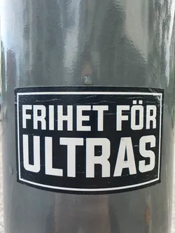 A black and white sticker with the text "FRIHET FÖR ULTRAS" on a gray surface. The sticker shows some wear and tear.