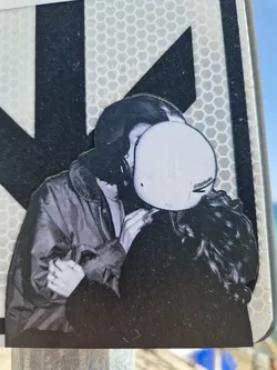 A black and white sticker depicting a couple sharing a tender kiss. The person on the left appears to be wearing a jacket, while the person on the right has long hair and is partially obscured by a helmet. The style is reminiscent of street photography, with a focus on intimacy and emotion.  The sticker is affixed to a reflective surface.
