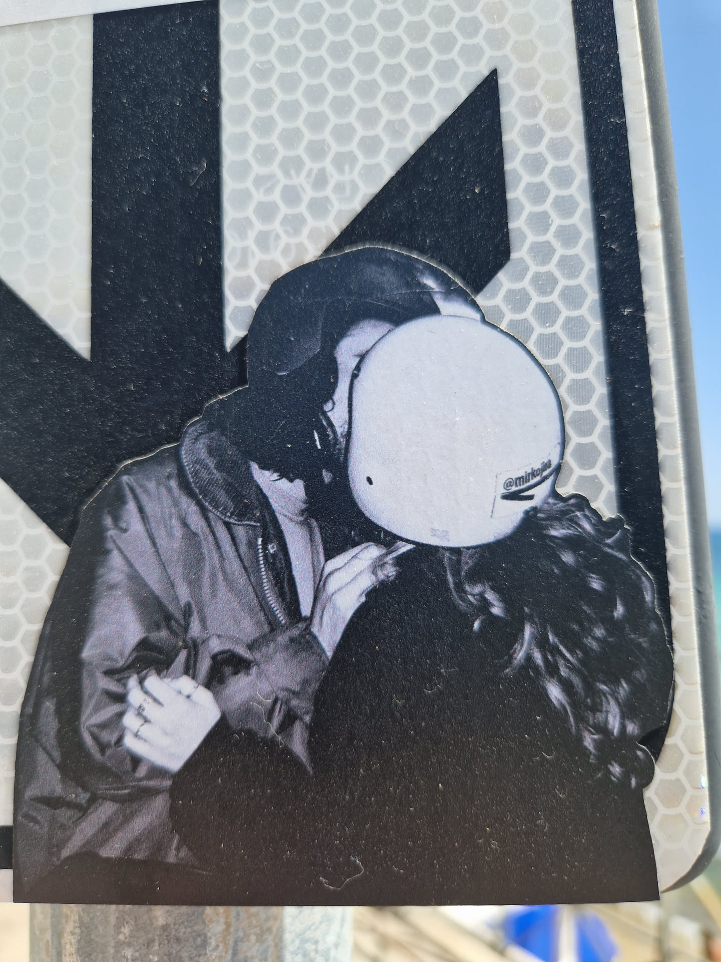A black and white sticker depicting a couple sharing a tender kiss. The person on the left appears to be wearing a jacket, while the person on the right has long hair and is partially obscured by a helmet. The style is reminiscent of street photography, with a focus on intimacy and emotion.  The sticker is affixed to a reflective surface.