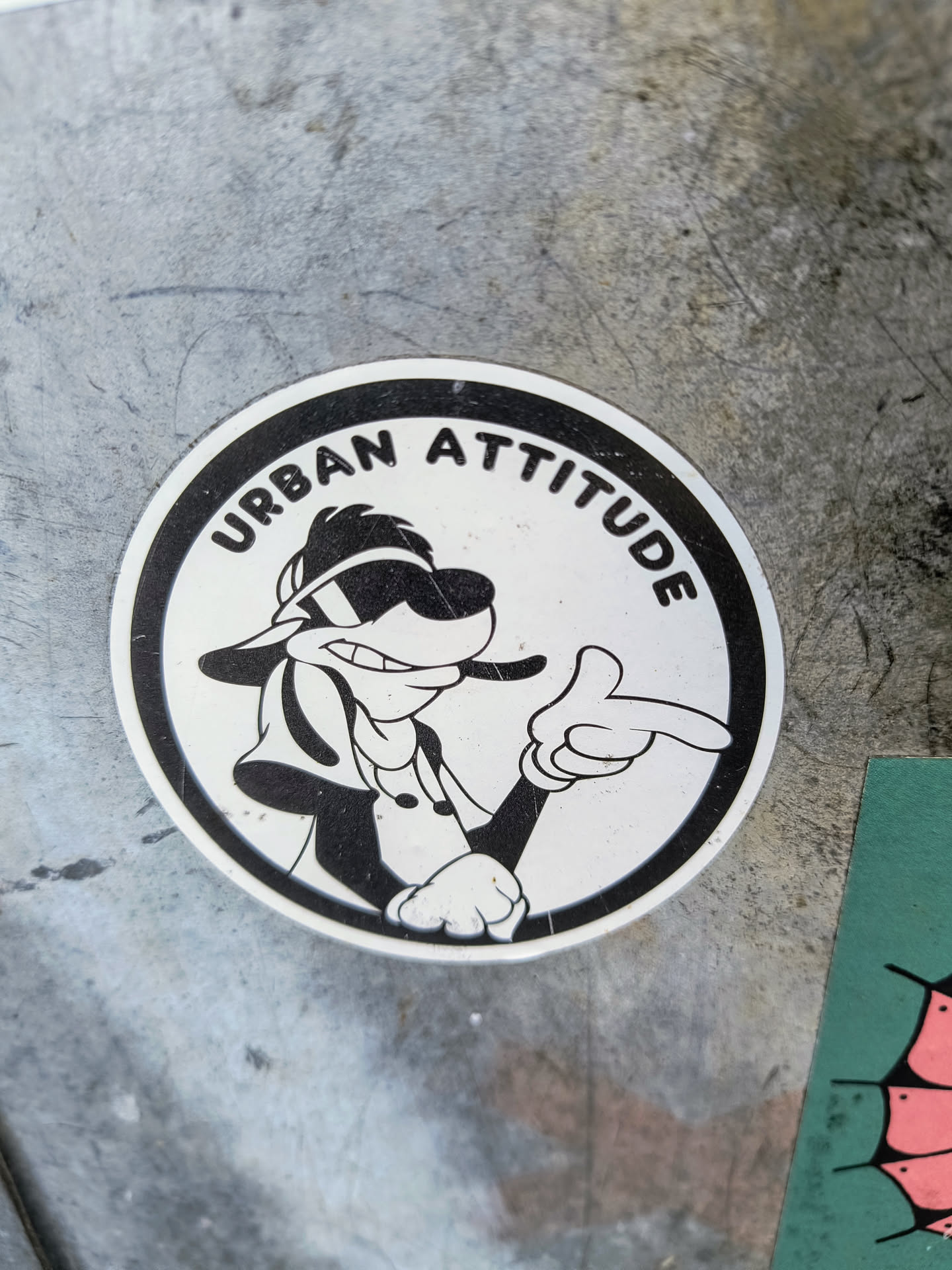 The sticker is circular and features a black and white cartoon image of a dog with sunglasses giving a pointing gesture. The words "Urban Attitude" are printed around the top edge of the circle. The sticker is on a metallic surface.