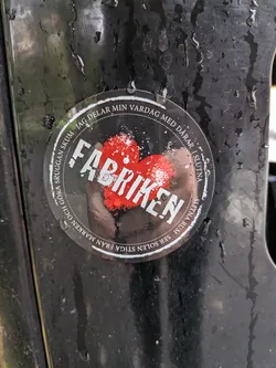 The sticker is circular and appears to be made of a transparent material, with some scuffing and water droplets on its surface. At the center of the sticker is a stylized red heart, rendered with a distressed, almost spray-painted texture, accented with white speckles. The word 'FABRIKEN' is prominently displayed across the heart in bold, white, sans-serif capital letters, also with a distressed appearance. Encircling the heart and the central text is a ring of black, with white text forming a motto. The visible text, read in a clockwise direction, includes fragments like 'JAG DELAR MIN VÅRDAG MED DARAR I SLUTNA', 'SLITNA RUM SER SOLEN STIGA FRÅN MARKEN OCH GÖRA SKUGGAN SKUM'. The overall aesthetic is rugged and perhaps industrial.