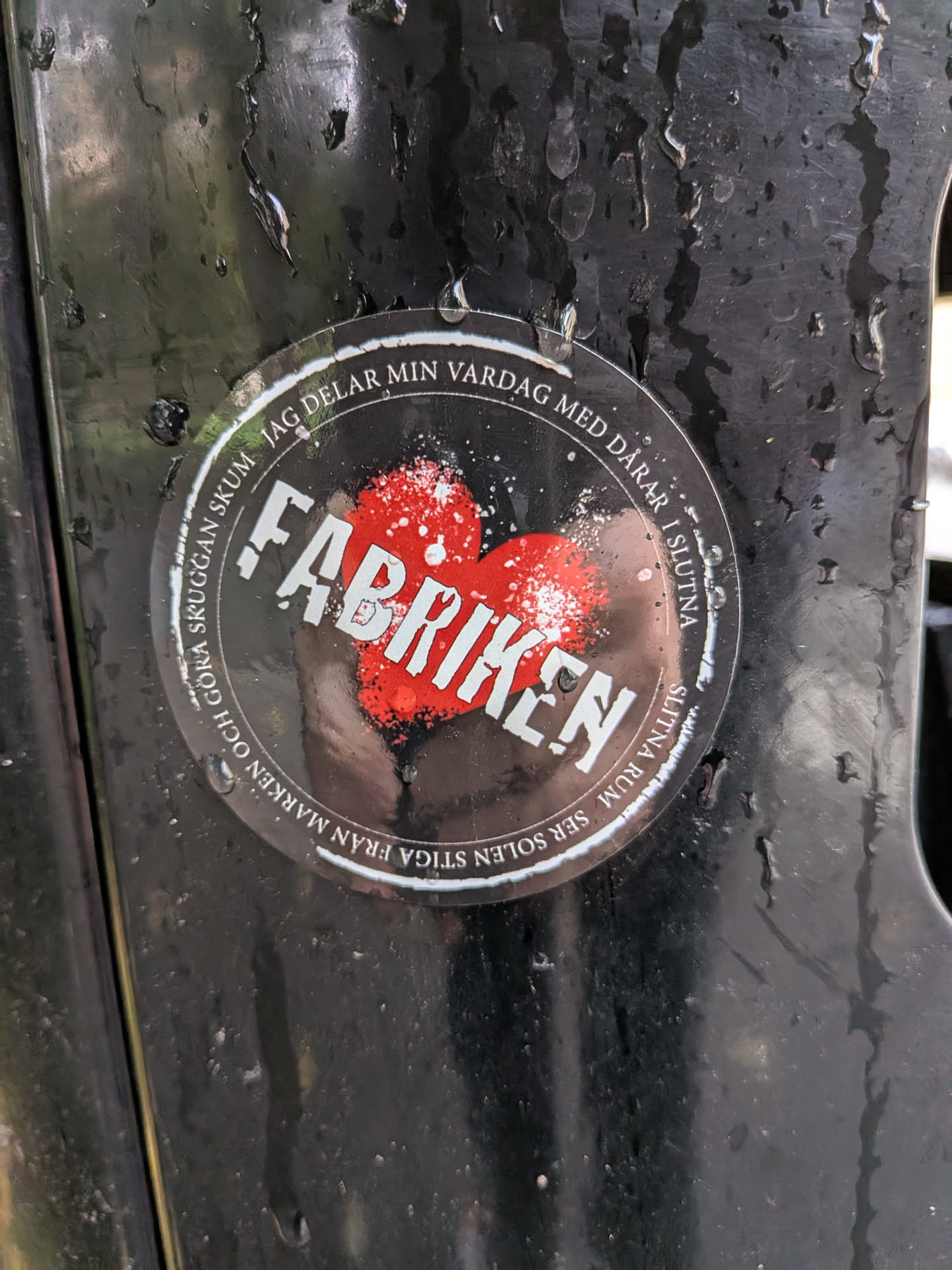 The sticker is circular and appears to be made of a transparent material, with some scuffing and water droplets on its surface. At the center of the sticker is a stylized red heart, rendered with a distressed, almost spray-painted texture, accented with white speckles. The word 'FABRIKEN' is prominently displayed across the heart in bold, white, sans-serif capital letters, also with a distressed appearance. Encircling the heart and the central text is a ring of black, with white text forming a motto. The visible text, read in a clockwise direction, includes fragments like 'JAG DELAR MIN VÅRDAG MED DARAR I SLUTNA', 'SLITNA RUM SER SOLEN STIGA FRÅN MARKEN OCH GÖRA SKUGGAN SKUM'. The overall aesthetic is rugged and perhaps industrial.