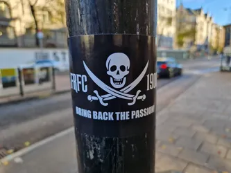 A black and white sticker with a pirate skull and crossbones design.  The text "RFC 1906" is visible on either side of the skull, and at the bottom, it reads "BRING BACK THE PASSION!". The sticker is affixed to a dark-colored pole on a city street.