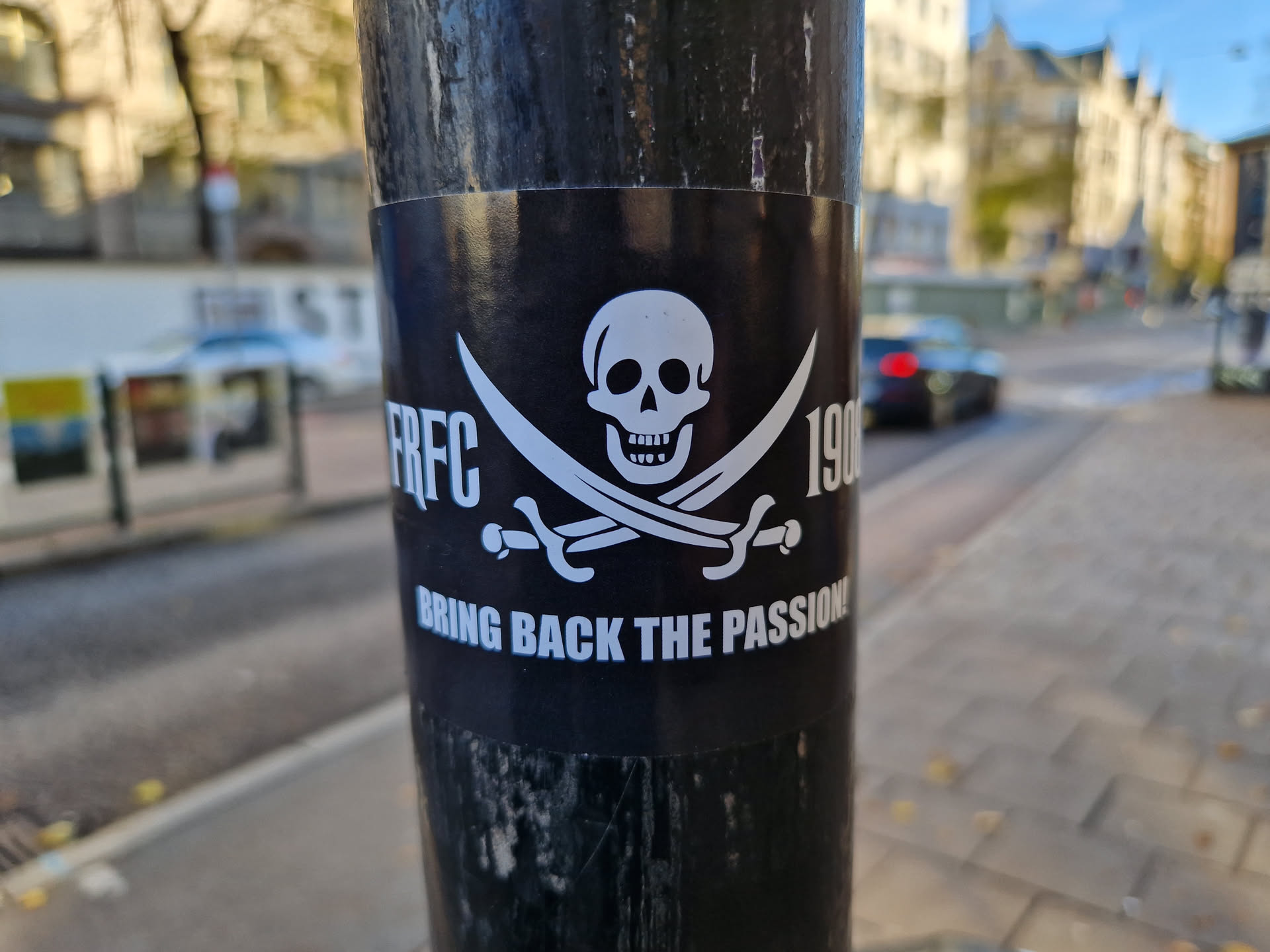 A black and white sticker with a pirate skull and crossbones design.  The text "RFC 1906" is visible on either side of the skull, and at the bottom, it reads "BRING BACK THE PASSION!". The sticker is affixed to a dark-colored pole on a city street.