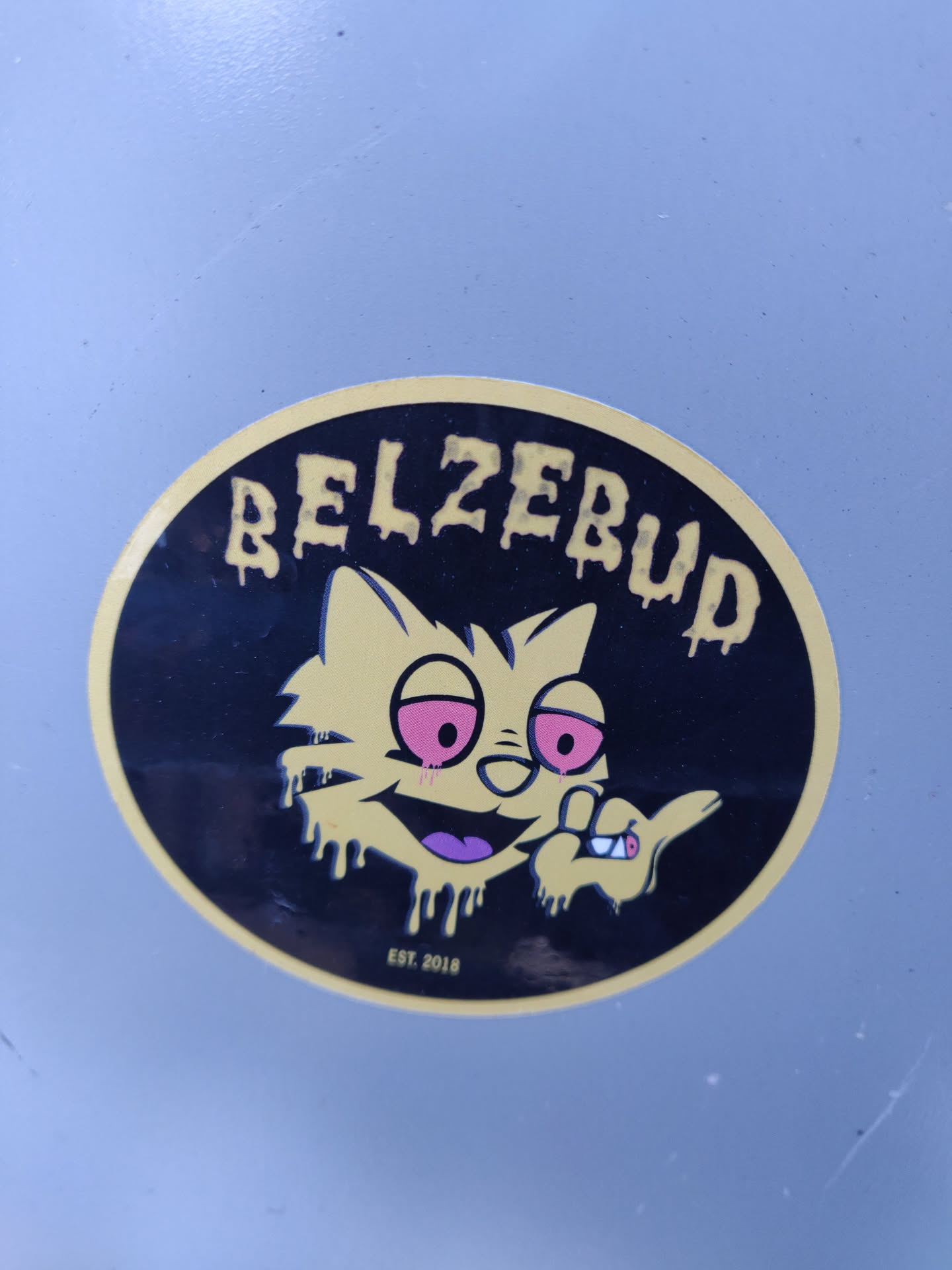 Round sticker featuring a cartoon cat with red eyes, holding a cigarette, with dripping paint effect. The text "Belzebud" is written in a dripping style above the cat. The text "EST. 2018" is written at the bottom.