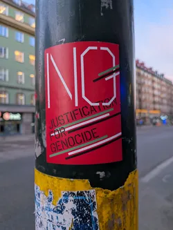 The sticker is predominantly red with large white lettering spelling out 'NG'. Below this, in smaller, stencil-like green lettering, it reads 'JUSTIFICATION FOR GENOCIDE'. The text is overlaid with thin, diagonal stripes of green, black, and white. The sticker appears to be adhered to a dark pole, with some wear and tear visible around its edges.