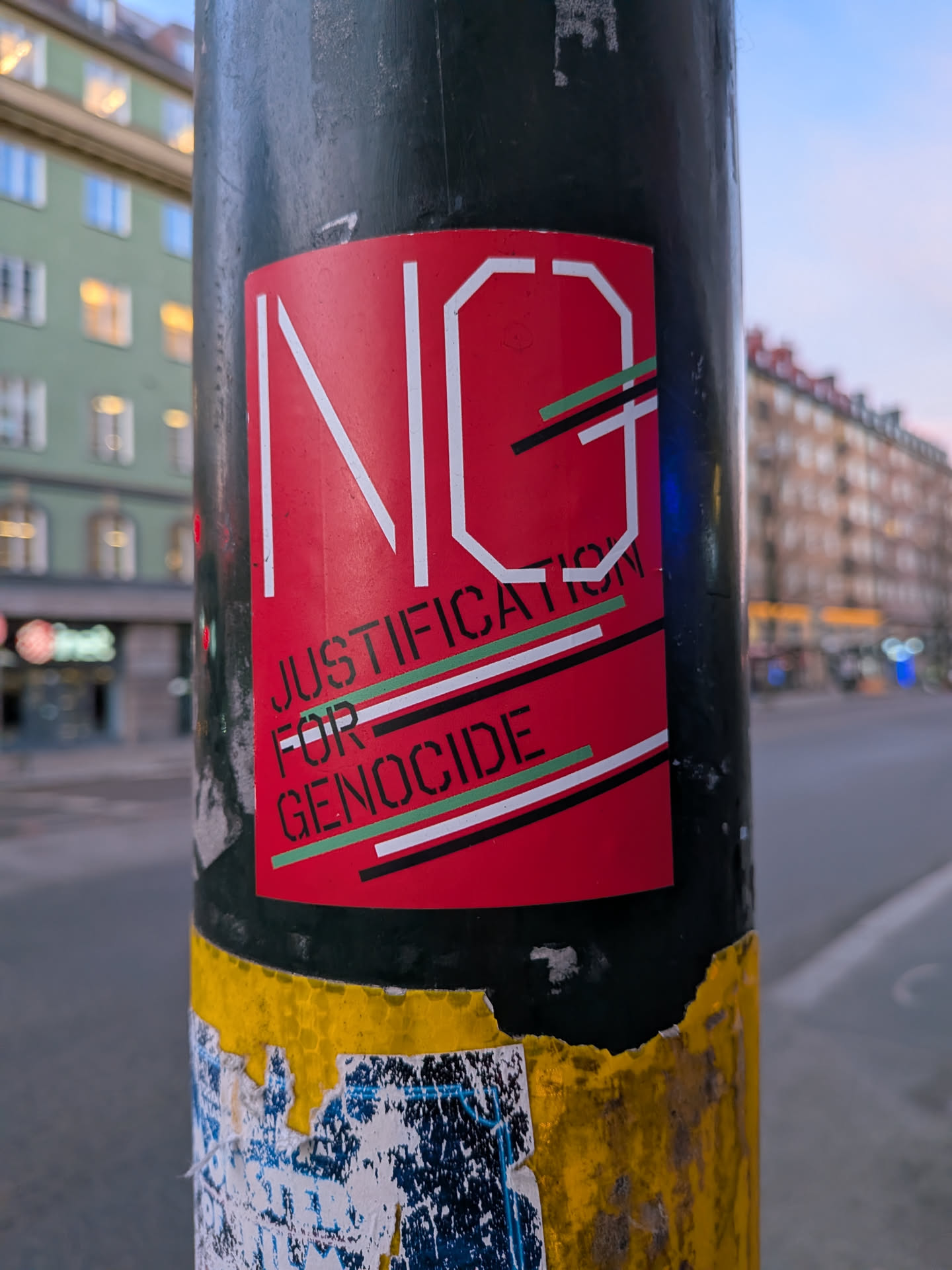 The sticker is predominantly red with large white lettering spelling out 'NG'. Below this, in smaller, stencil-like green lettering, it reads 'JUSTIFICATION FOR GENOCIDE'. The text is overlaid with thin, diagonal stripes of green, black, and white. The sticker appears to be adhered to a dark pole, with some wear and tear visible around its edges.
