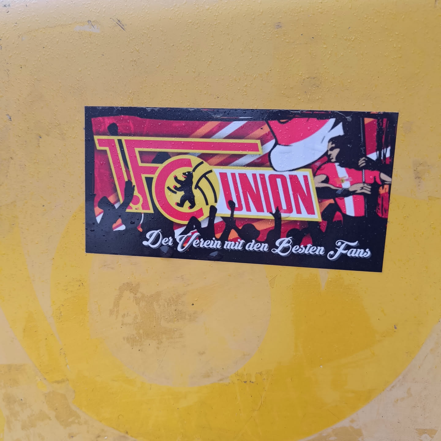 A sticker featuring the logo of 1. FC Union Berlin, a German football club. The text on the sticker translates to “the club with the best fans”.  The design includes silhouettes of cheering fans and a stylized image of a player.