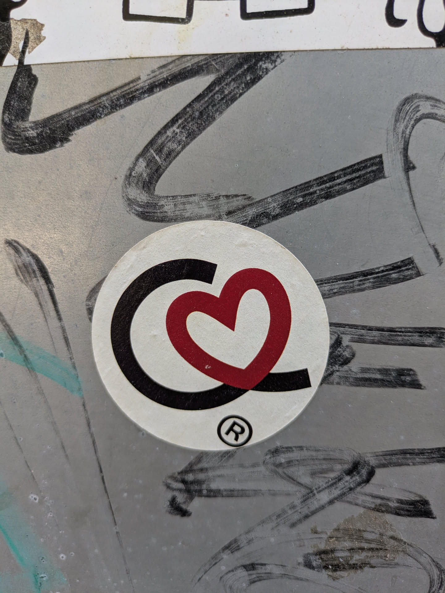 This sticker features a black 'C' shape embracing a red heart, creating a visually appealing logo. The sticker is circular and appears to be adhered to a textured, potentially metallic surface, adorned with other markings.
