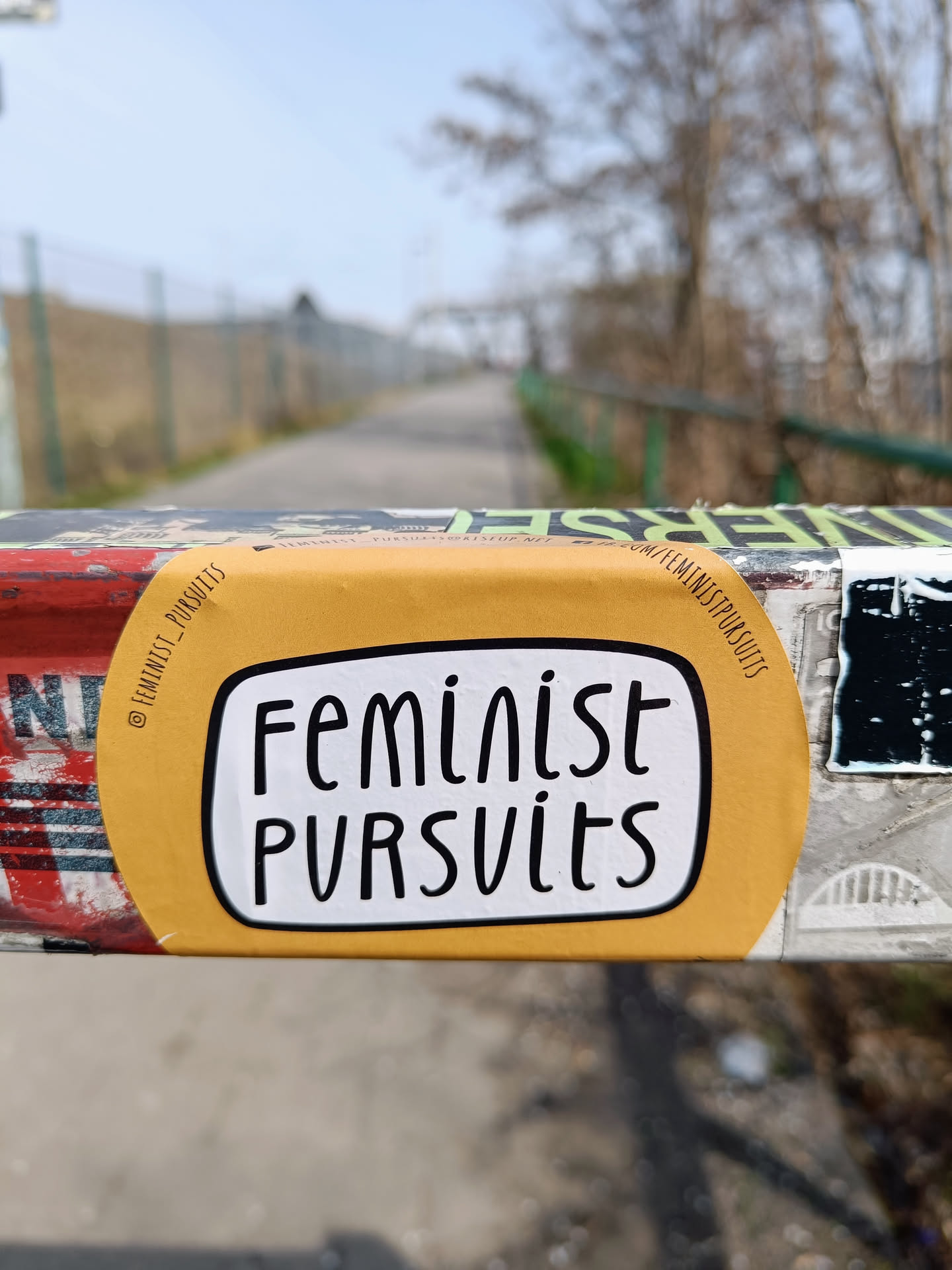 A yellow sticker with the text "Feminist Pursuits" written in a stylized font. The sticker is affixed to a metal surface outdoors.
