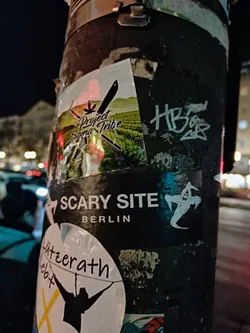 A sticker featuring the logo for Project Soma Tribe, a company or group related to cannabis, is seen on a pole in Berlin. The sticker has a mountainous landscape and a stylized cannabis leaf in its design. Other stickers are present on the same pole, including one that says "Scary Site Berlin."