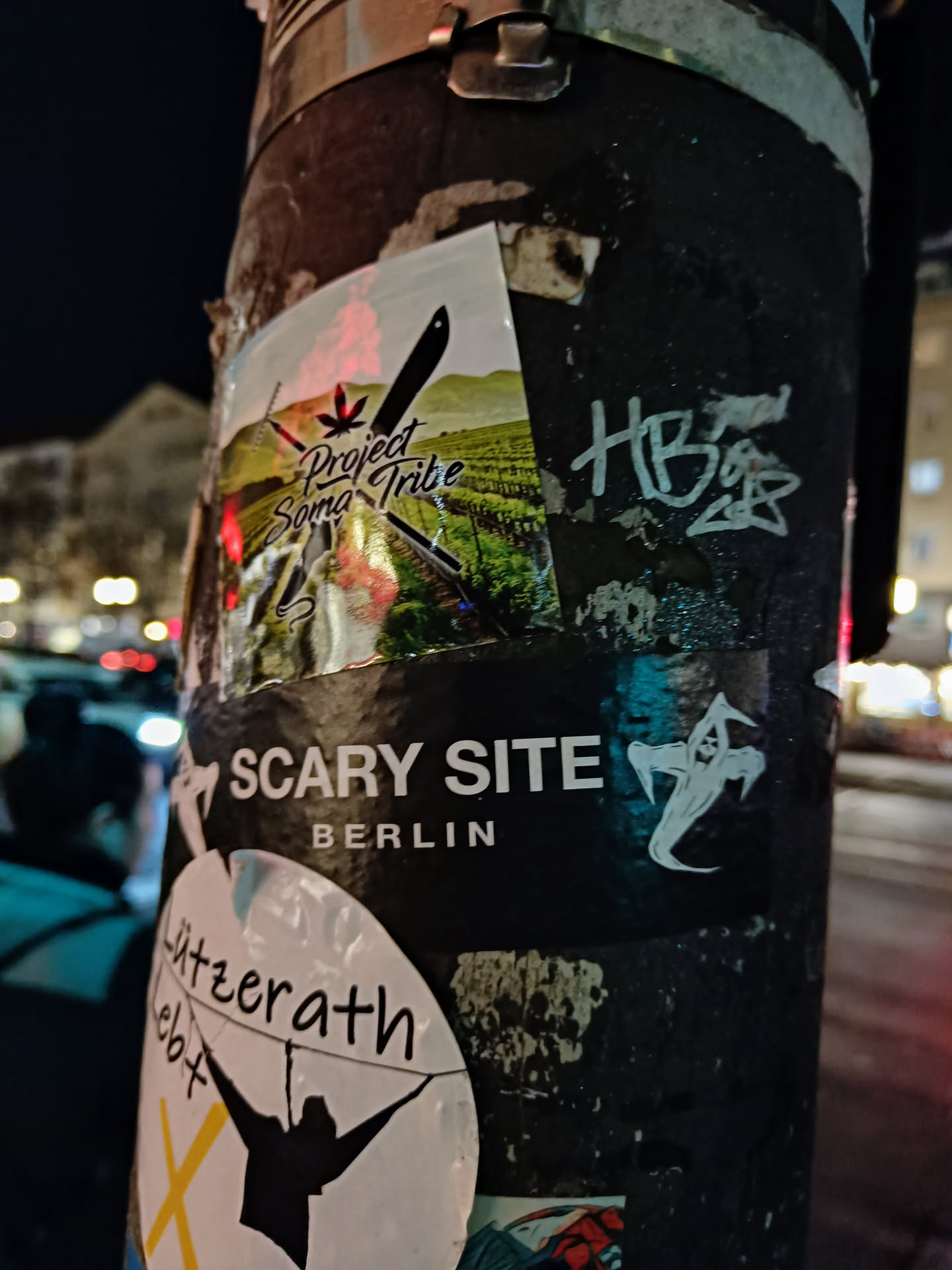 A sticker featuring the logo for Project Soma Tribe, a company or group related to cannabis, is seen on a pole in Berlin. The sticker has a mountainous landscape and a stylized cannabis leaf in its design. Other stickers are present on the same pole, including one that says "Scary Site Berlin."