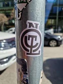 A black and white circular sticker with a stylized logo featuring the letters 'NT' above a symbol that looks like a stylized 'ψ' (psi). The sticker is affixed to a metal pole, which is also covered in other stickers. The background shows a city street scene.