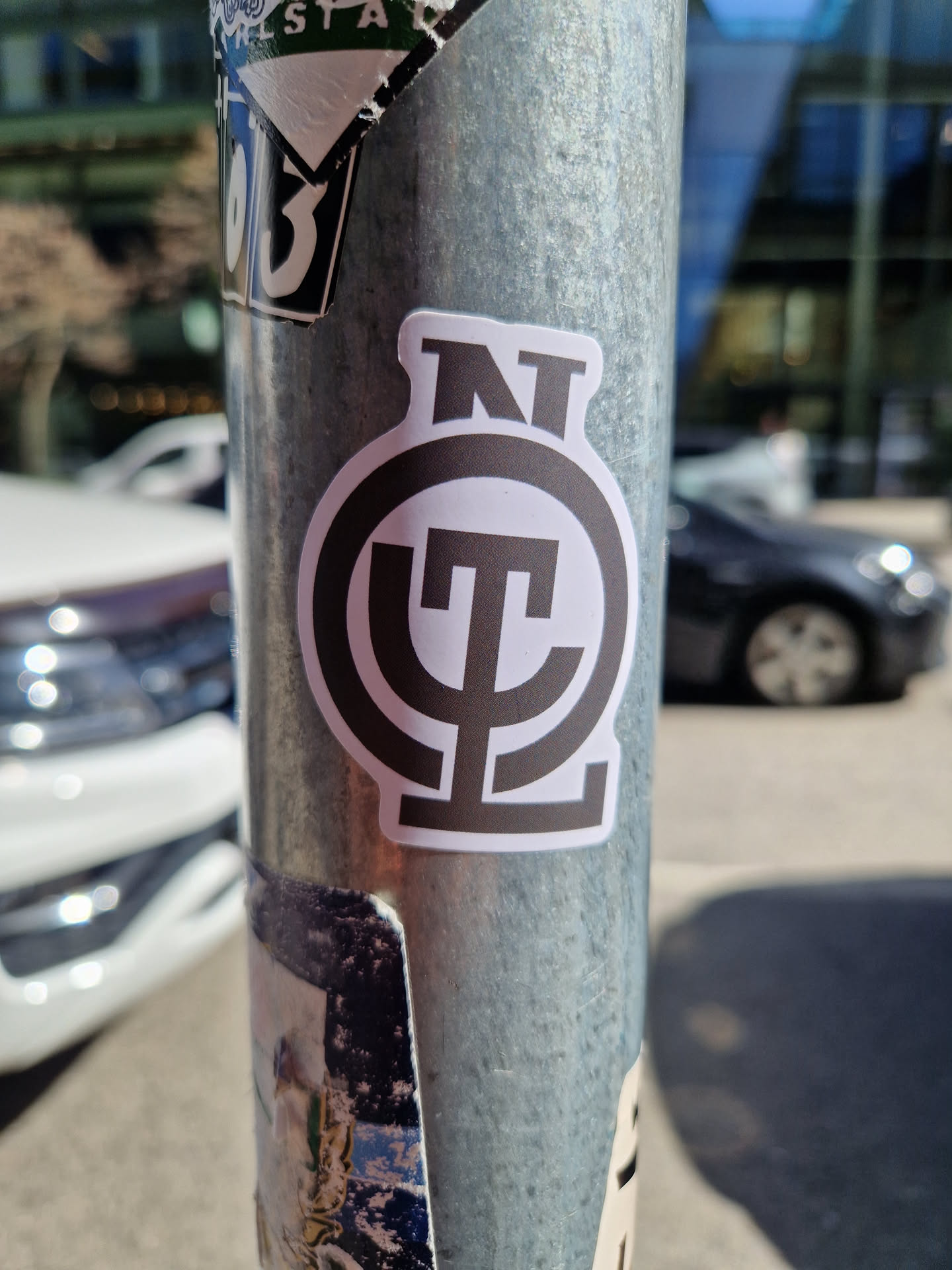 A black and white circular sticker with a stylized logo featuring the letters 'NT' above a symbol that looks like a stylized 'ψ' (psi). The sticker is affixed to a metal pole, which is also covered in other stickers. The background shows a city street scene.