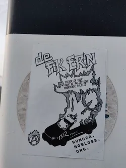Black and white sticker with a drawing of a burning police car and the text "De fik erin" which translates to "Set it on fire". The sticker also includes the text "Stenen en vuur voor de racistische moordende politie", which translates to "Stones and fire for the racist murdering police". At the bottom of the sticker is the text "Rumor Noblogs.org".  The anarchist symbol is also present on the bottom left.