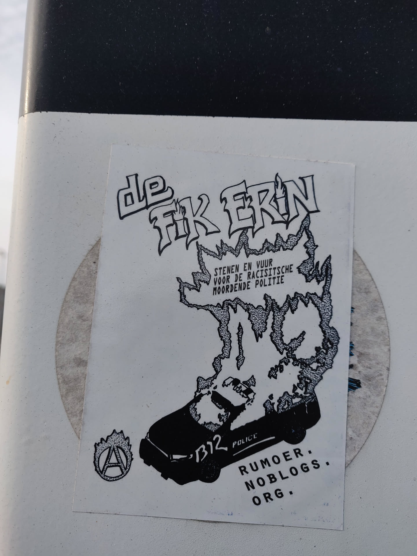 Black and white sticker with a drawing of a burning police car and the text "De fik erin" which translates to "Set it on fire". The sticker also includes the text "Stenen en vuur voor de racistische moordende politie", which translates to "Stones and fire for the racist murdering police". At the bottom of the sticker is the text "Rumor Noblogs.org".  The anarchist symbol is also present on the bottom left.