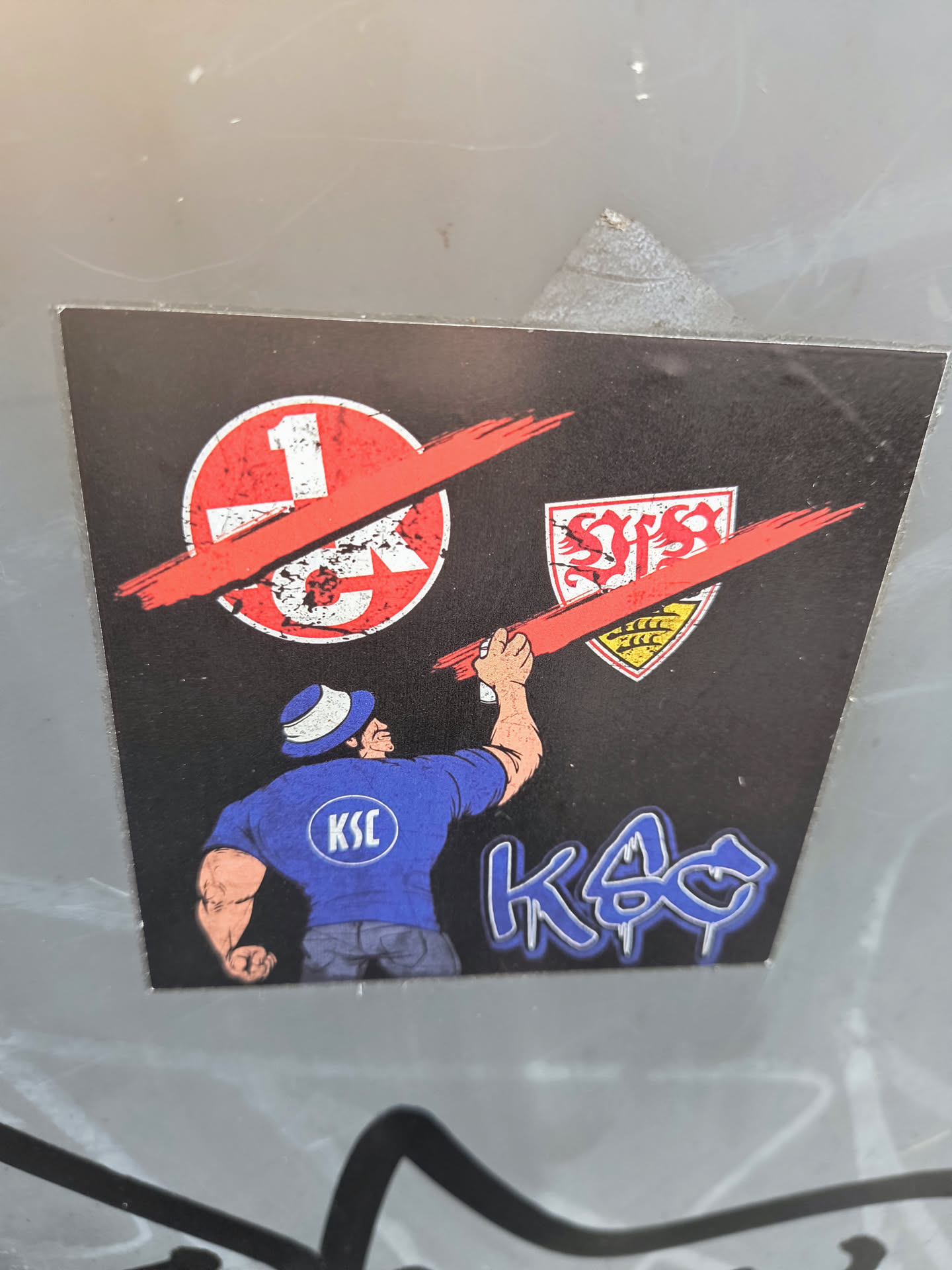 The sticker features a man in a blue shirt with 'KSC' on the back, who is using a red spray can to paint over the logos of 1. FC Kaiserslautern and VfB Stuttgart. This symbolizes rivalry between the teams.