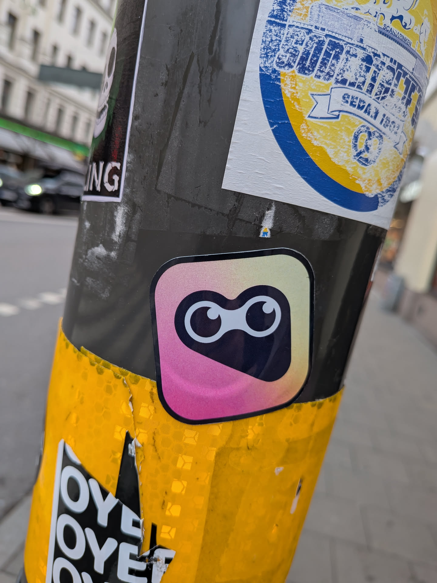 The sticker is centrally located and features a rounded square shape with a gradient background transitioning from pink to yellow and then to a light blue. Within this shape, there are two large, circular eyes with black pupils and white irises, giving it a cartoonish and friendly appearance. The sticker has a subtle border.