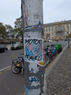 Street sticker A square white sticker featuring bold, stylized black lettering reading 'MOTOR' across the top and 'TRAVEL' along the bottom. The center features a blue triangular emblem with the text 'RKS MOTOR' and a pink graphic, adjacent to a detailed compass rose rendered in blue, red, and black. Palm tree silhouettes in light blue accent the corners, all adhered to a weathered urban metal pole.