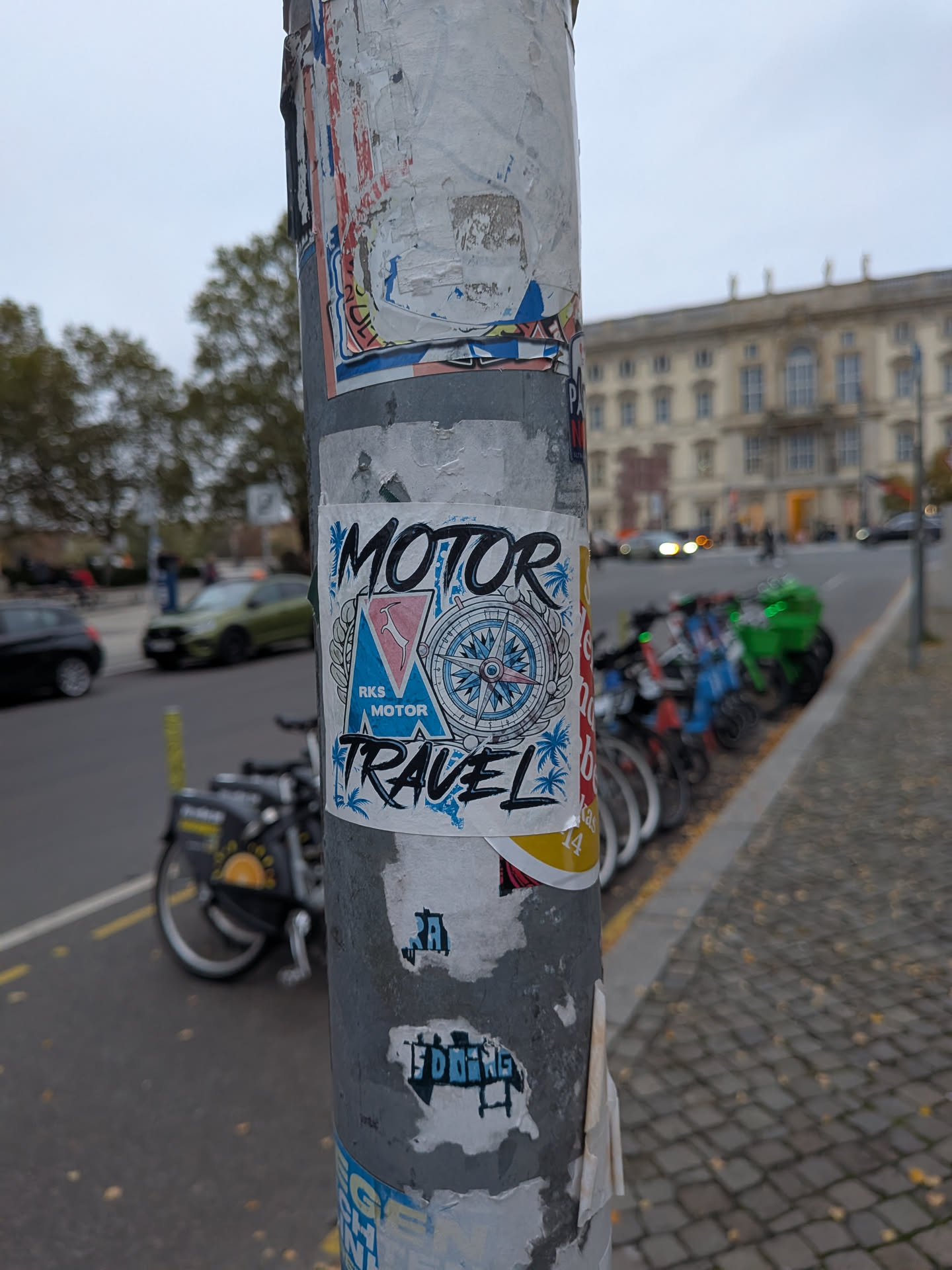 A square white sticker featuring bold, stylized black lettering reading 'MOTOR' across the top and 'TRAVEL' along the bottom. The center features a blue triangular emblem with the text 'RKS MOTOR' and a pink graphic, adjacent to a detailed compass rose rendered in blue, red, and black. Palm tree silhouettes in light blue accent the corners, all adhered to a weathered urban metal pole.