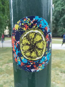 This round sticker showcases a detailed illustration of a lemon slice in yellow and black, overlaid on a dynamic backdrop of blue, red, and orange paint splatters. The style is reminiscent of pop art or graffiti.