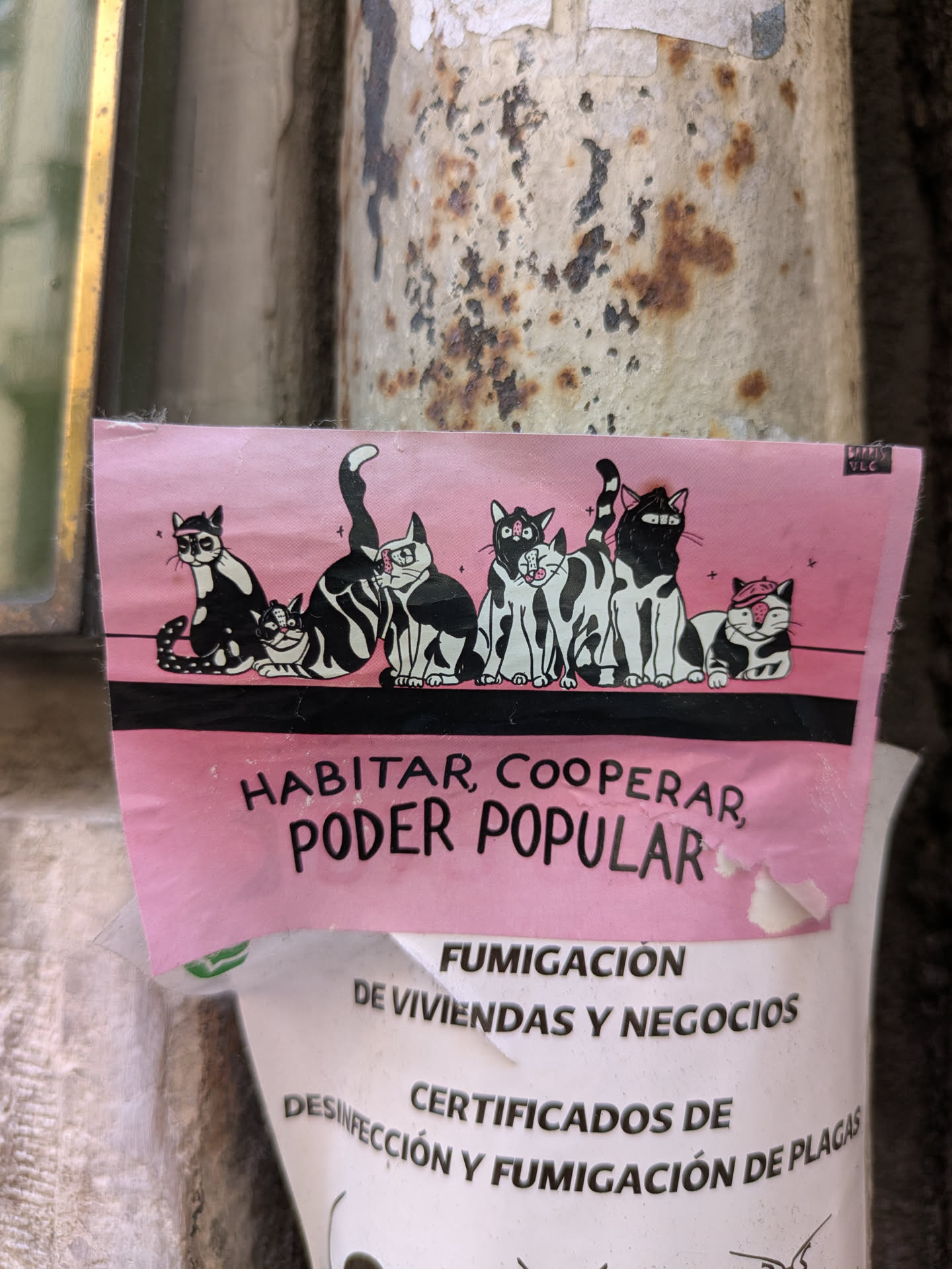 The pink sticker shows a row of black and white cartoon cats. Below the cats, the text reads "HABITAR, COOPERAR, PODER POPULAR".  The sticker appears to be on a pole or wall, partially covering another sticker.