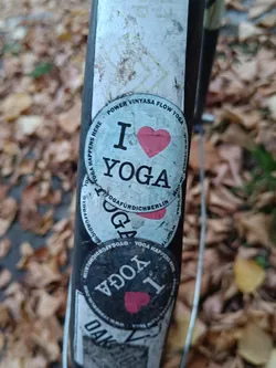 A round sticker with the text "I ♥ YOGA" on it. There are other stickers on the same surface, which appears to be a bike. The sticker is slightly worn out and dirty. The background is blurry, but it looks like fallen leaves on the ground.