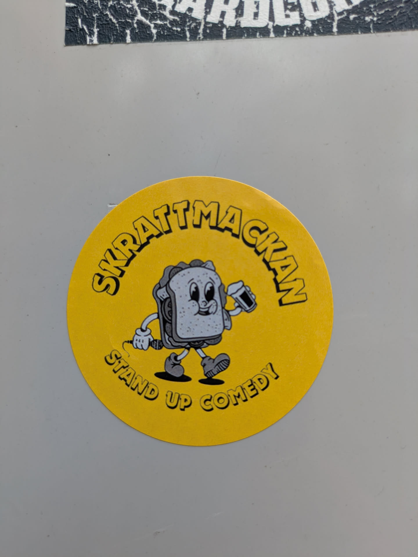 A round, yellow sticker featuring a sandwich cartoon character holding a microphone. The text around the character reads "SKRATTMACKAN STAND UP COMEDY".