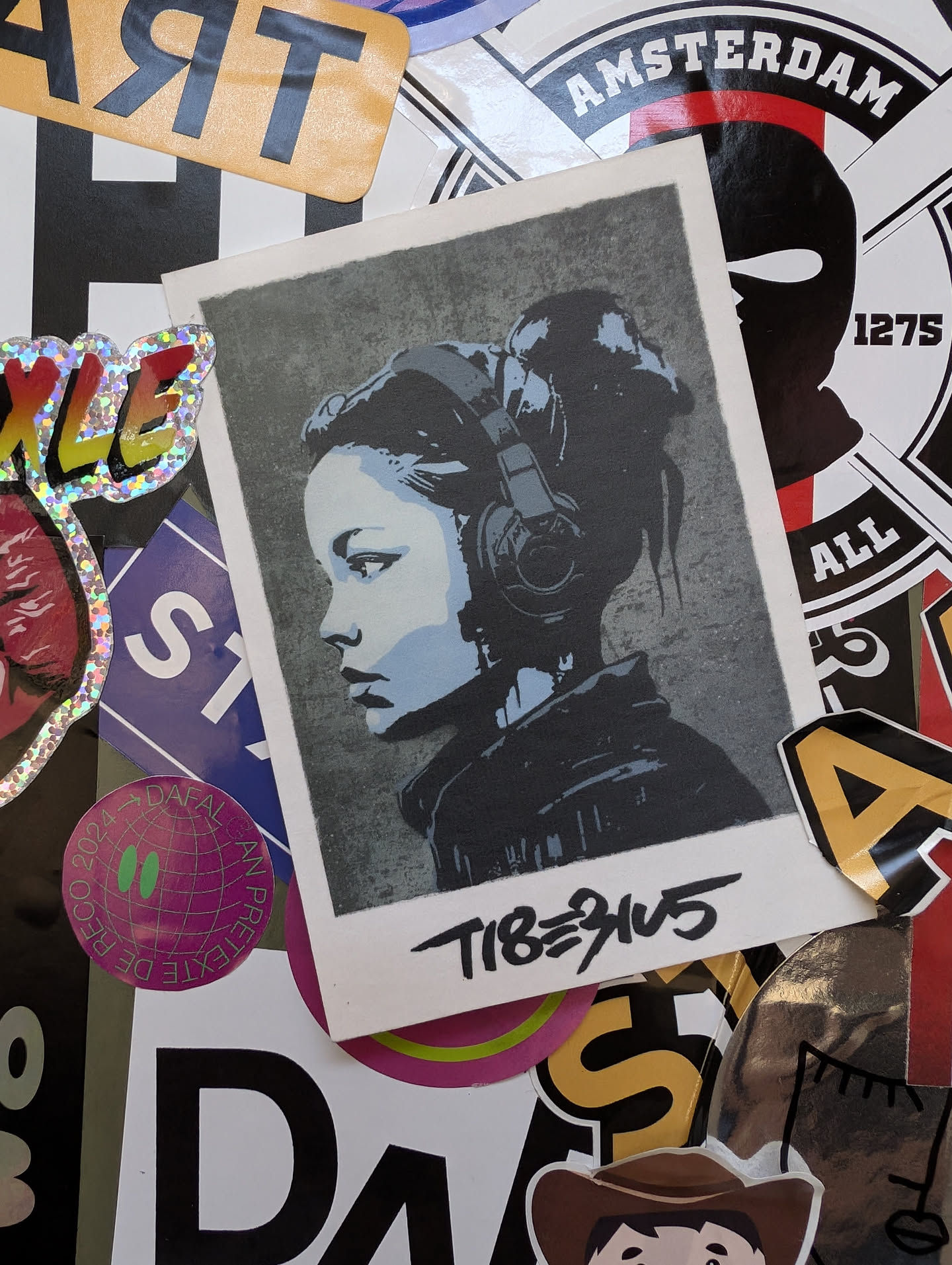 A sticker featuring a stylized portrait of a girl wearing headphones. The art style is reminiscent of street art or stencil work, with a slightly gritty texture. The sticker has the artist's tag 'TIB3945' written underneath the artwork.