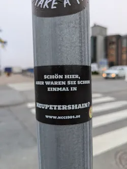 A black and white sticker on a metal pole with a German phrase.  It promotes a website related to Neupettershain.