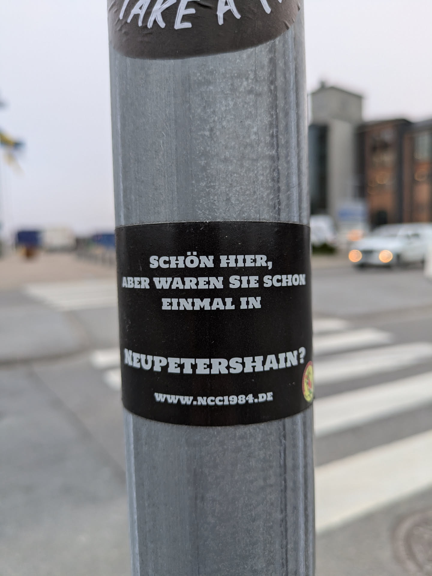 A black and white sticker on a metal pole with a German phrase.  It promotes a website related to Neupettershain.