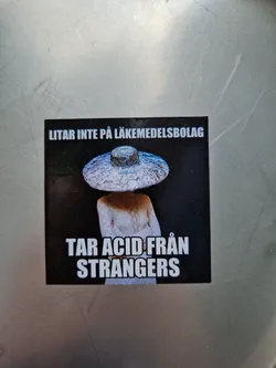 A sticker with a person wearing a large hat and the text "LITAR INTE PÅ LÄKEMEDELSBOLAG" (Swedish for "Don't trust pharmaceutical companies") and "TAR ACID FRÅN STRANGERS". The image is dark and mysterious.