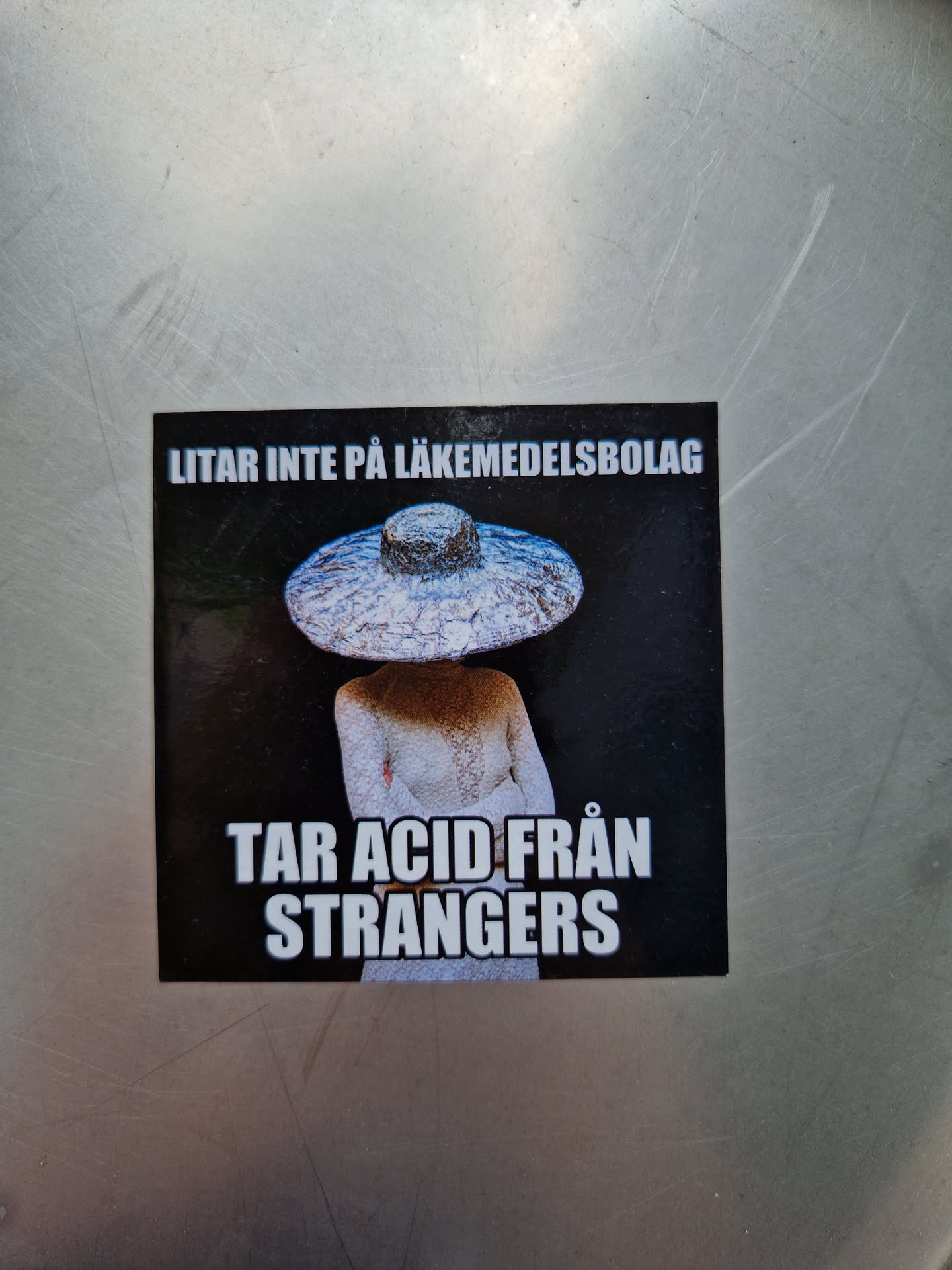 A sticker with a person wearing a large hat and the text "LITAR INTE PÅ LÄKEMEDELSBOLAG" (Swedish for "Don't trust pharmaceutical companies") and "TAR ACID FRÅN STRANGERS". The image is dark and mysterious.