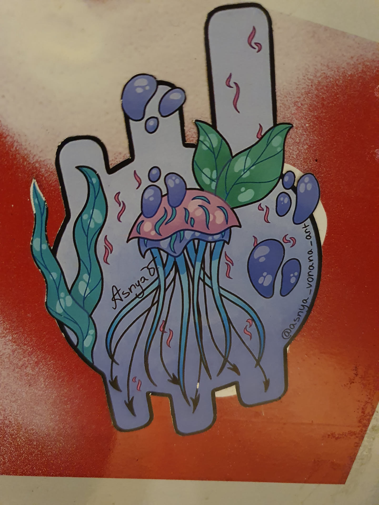 A hand-drawn style sticker featuring a jellyfish with seaweed and leaves. The jellyfish is primarily pink and purple with blue tentacles. The background is a light purple shape resembling a hand. The artist's signature and Instagram handle are visible in the artwork.