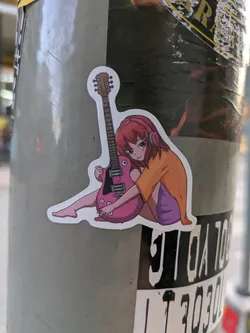 A sticker featuring an anime-style illustration of a girl with headphones sitting with a pink electric guitar. 