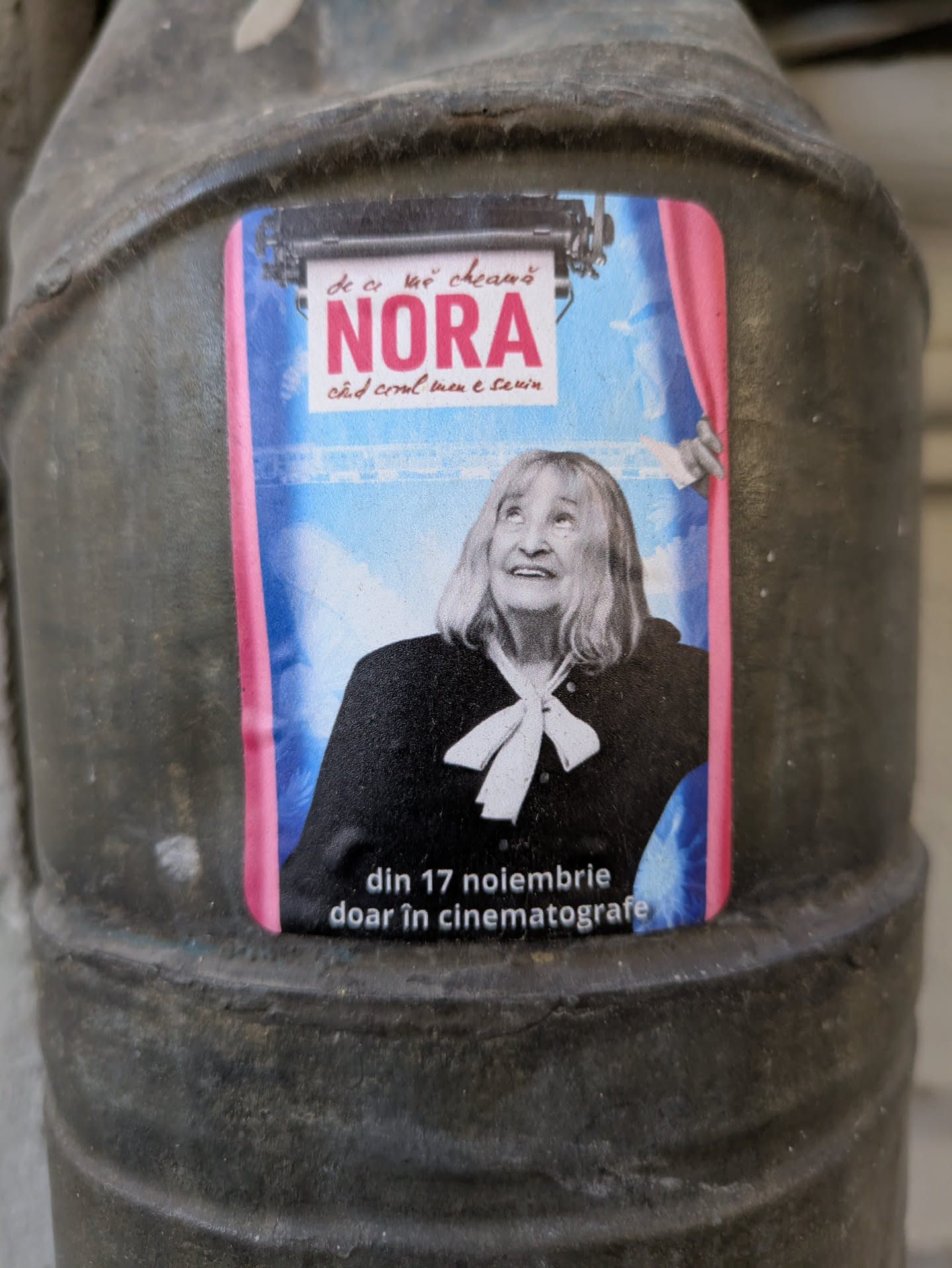 This is a promotional sticker for the movie "De ce mă cheamă Nora", featuring a woman in a black jacket and a white ribbon tied around her neck. The sticker advertises the release of the movie in cinemas starting November 17th. The sticker is pasted on a gray metal surface.
