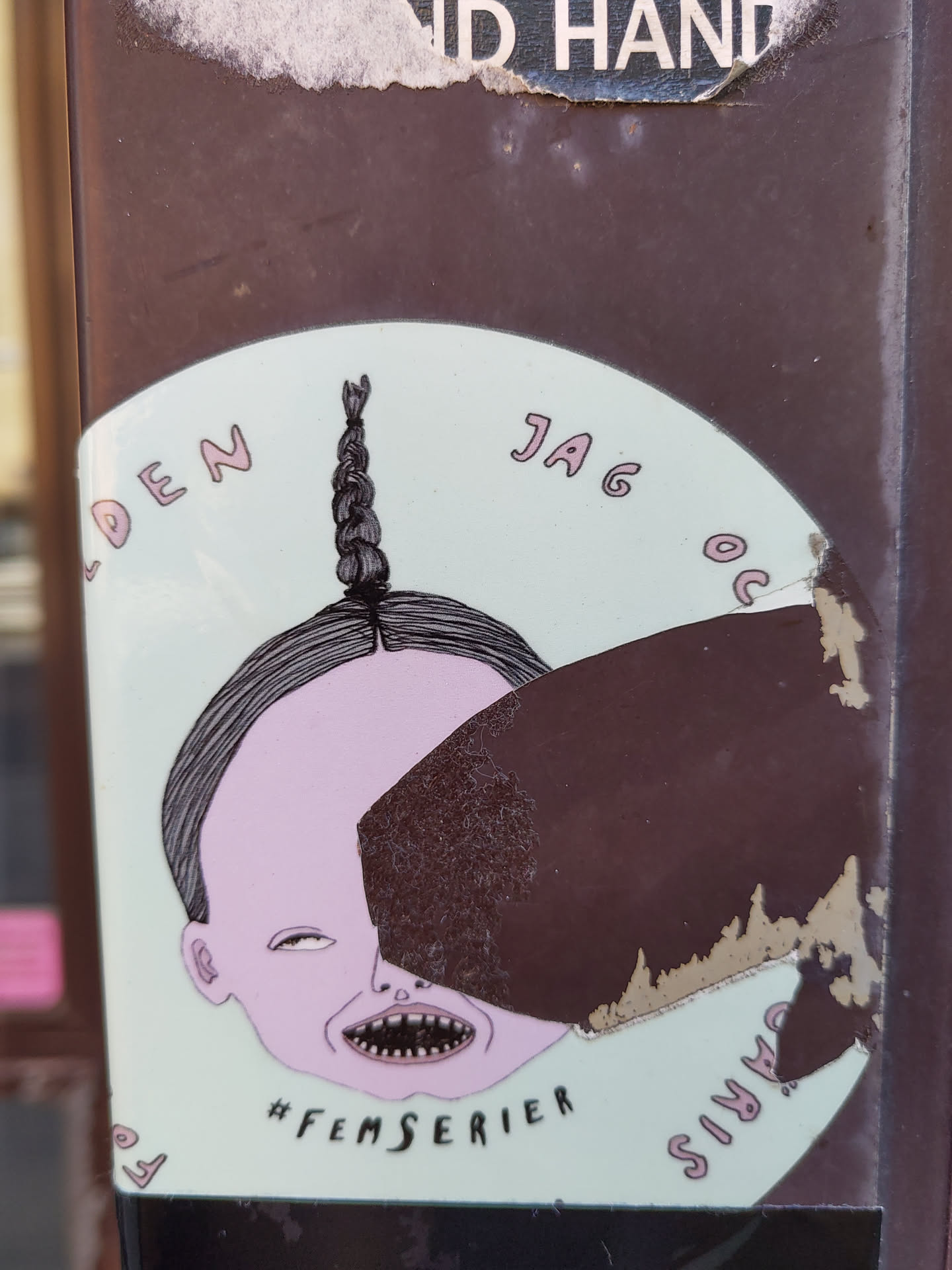 Round sticker with a pastel green background featuring a stylized illustration. A character's head with a braid is depicted, partially obscured by a dark brown irregular shape. Text in a pale pink font surrounds the illustration, and the hashtag #femserier is visible beneath it. The sticker shows some wear and tear, with edges slightly damaged.