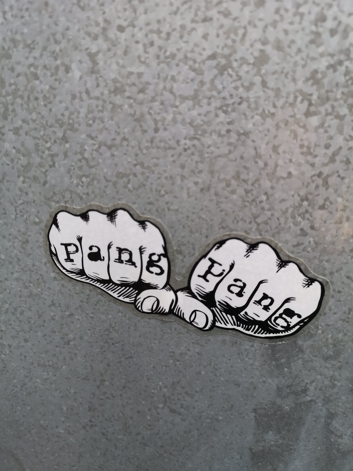 A black and white sticker featuring two clenched fists, side-by-side, with knuckles facing forward. Each fist has the word 'Pang' written across its knuckles in a bold, serif-style font. The fists are rendered in a graphic, illustrative style with thick black outlines and cross-hatching for shading, reminiscent of traditional tattoo art or comic book imagery. The sticker has a clear, die-cut border around the white design, making it stand out against the textured, speckled grey background, which appears to be galvanized metal.